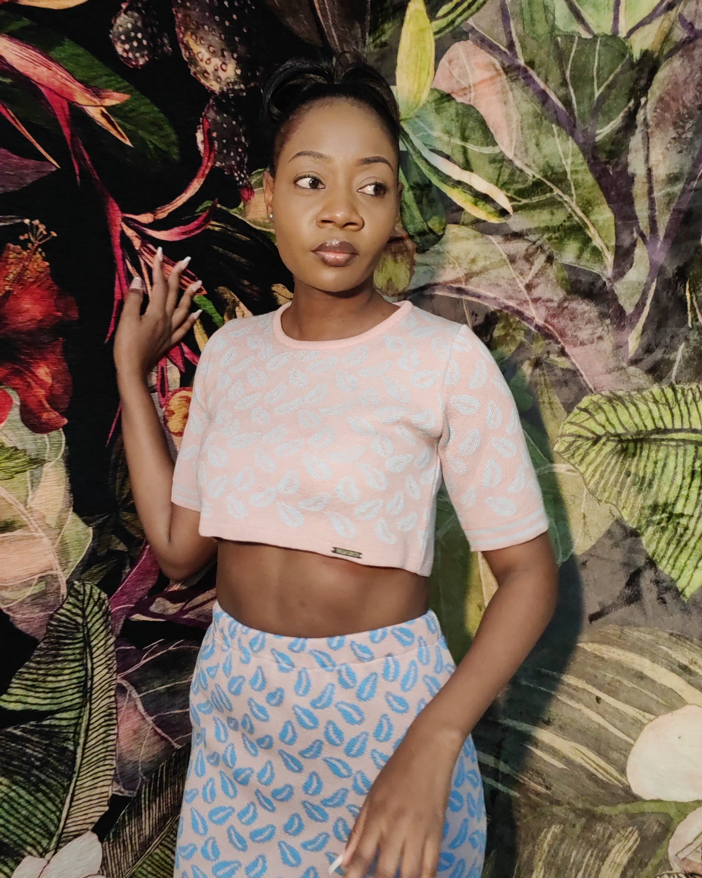 Woman in a pink top and blue patterned skirt standing against a tropical leaf background