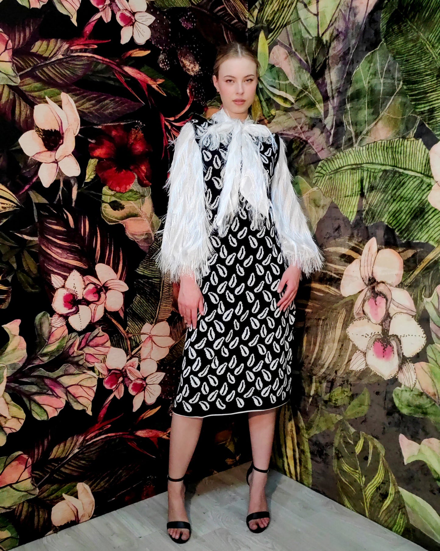 Woman in a black and white patterned dress with a white shawl standing against a floral and leaf-patterned wall.