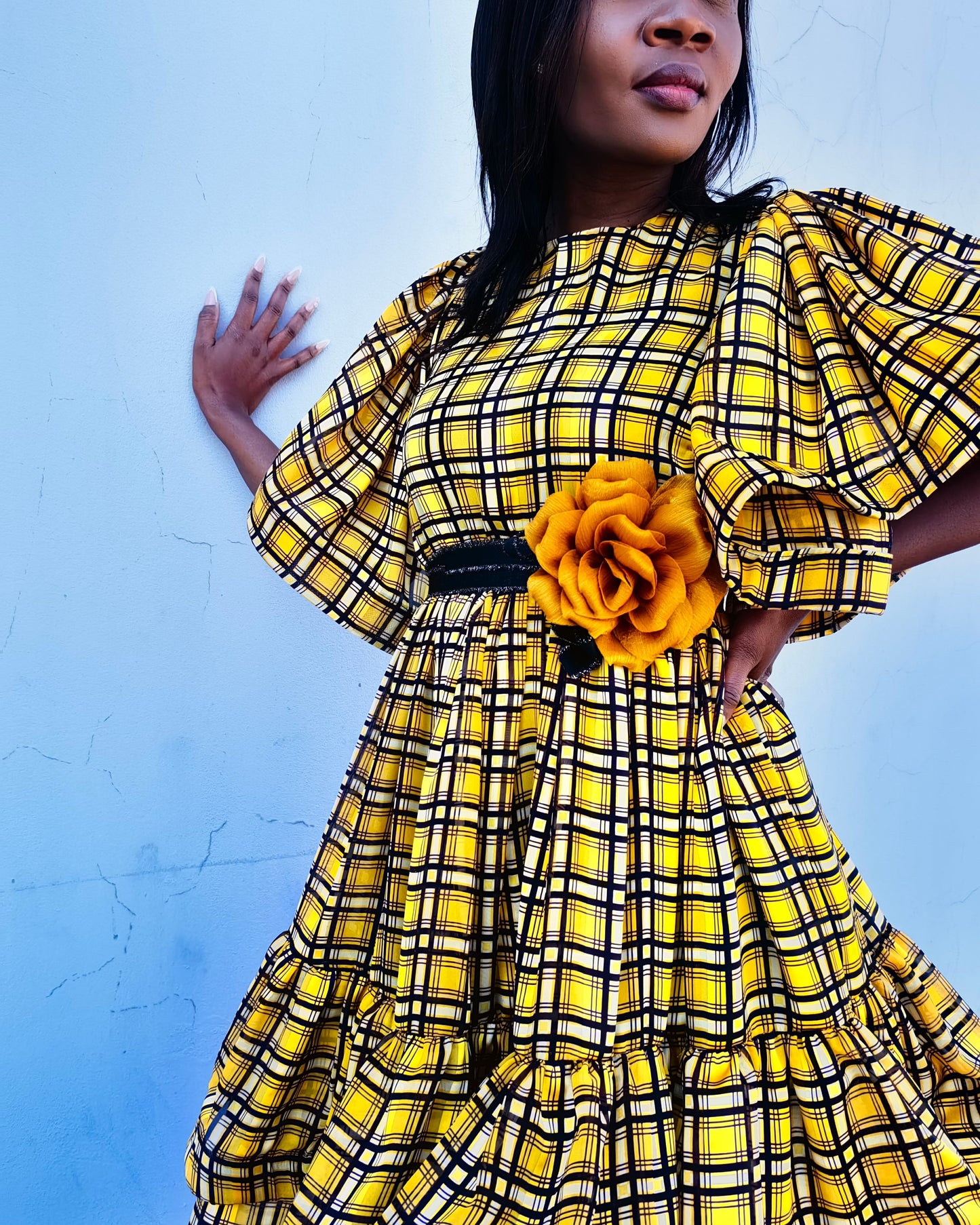 Woman wearing a yellow and black checkered dress with a large yellow flower accessory against a blue background