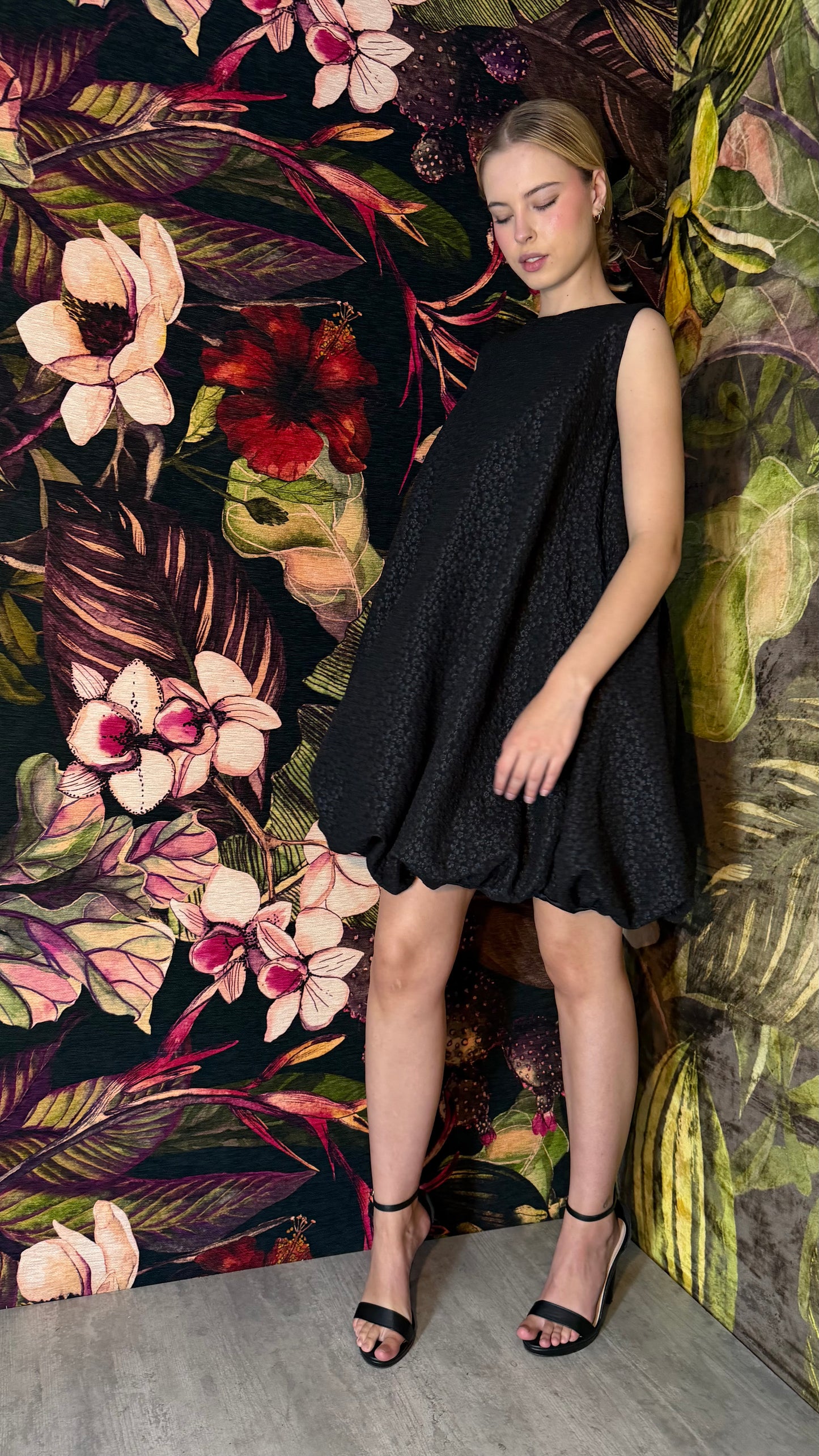 Woman in a black dress standing against a vibrant floral wall.