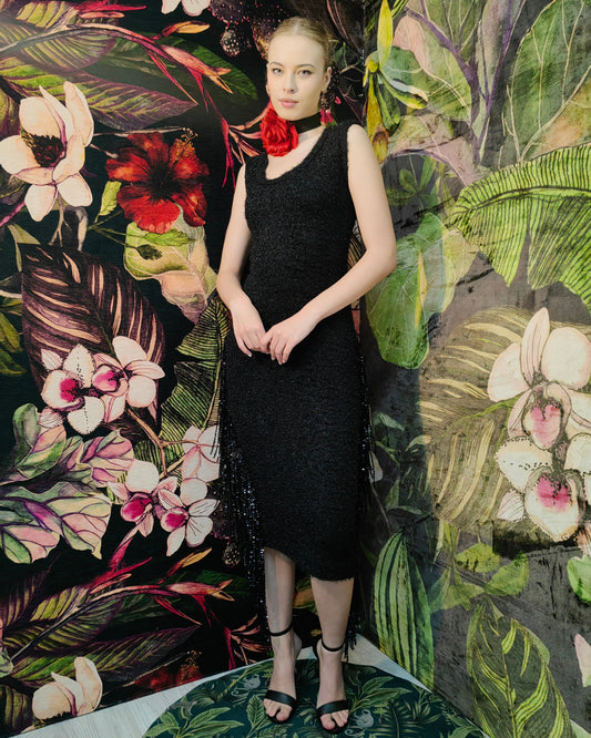 Woman in a black dress standing against a vibrant floral and leaf-patterned wall.
