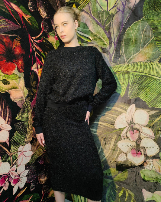 Woman in a black outfit standing against a colorful floral and leaf patterned wall.
