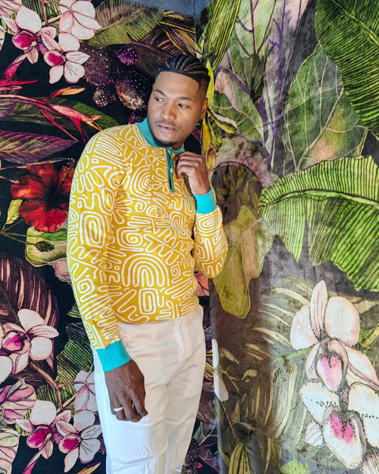Man wearing a yellow patterned shirt and beige pants standing against a colorful floral and leaf background