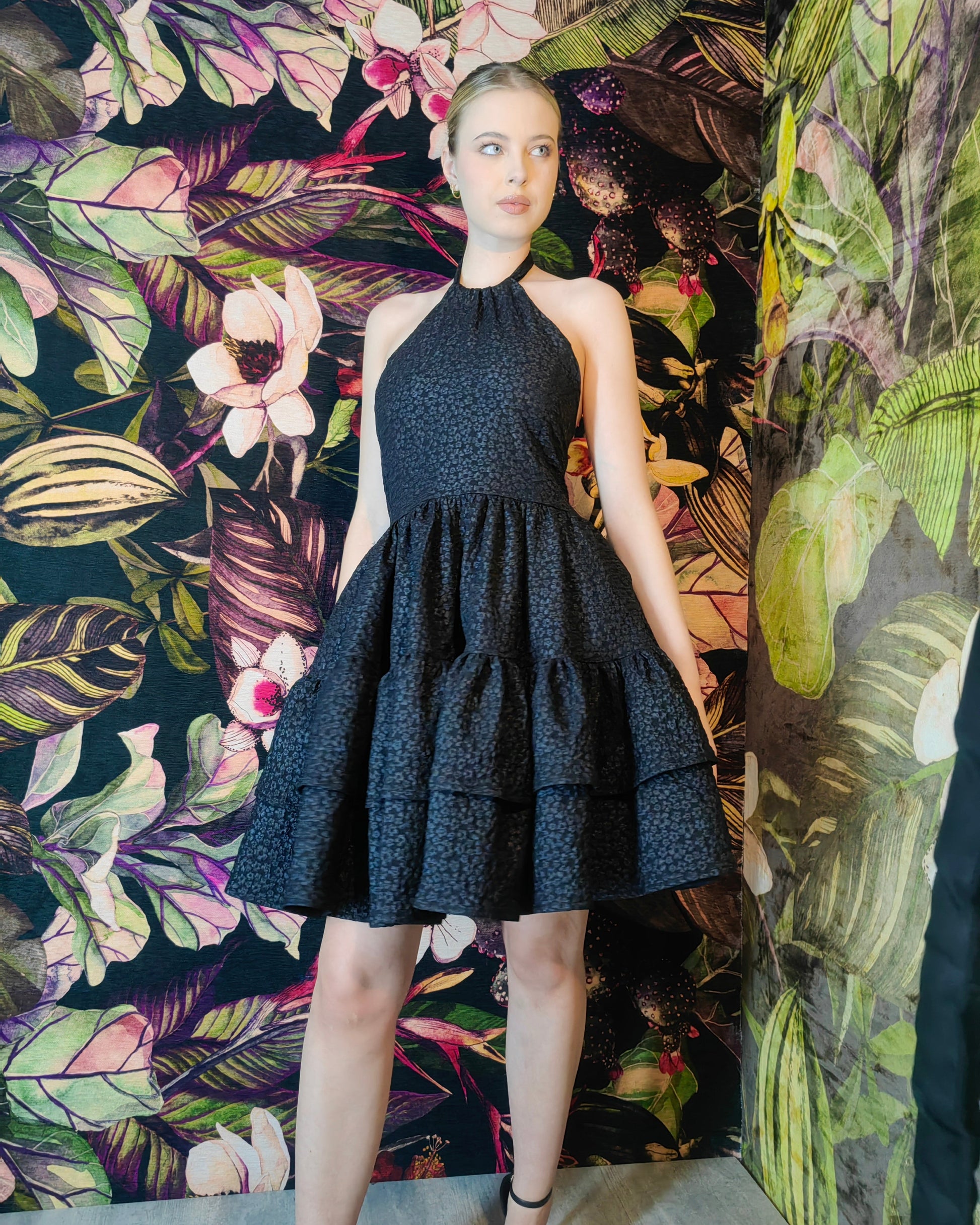 Woman wearing a black dress against a floral and leaf-patterned wall.
