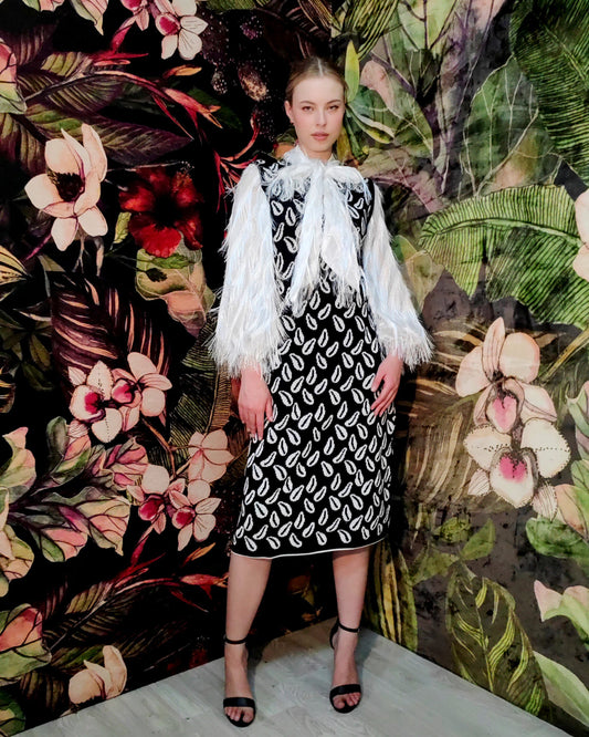 Woman in a black and white patterned dress with a white shawl standing against a floral and leaf-patterned wall.