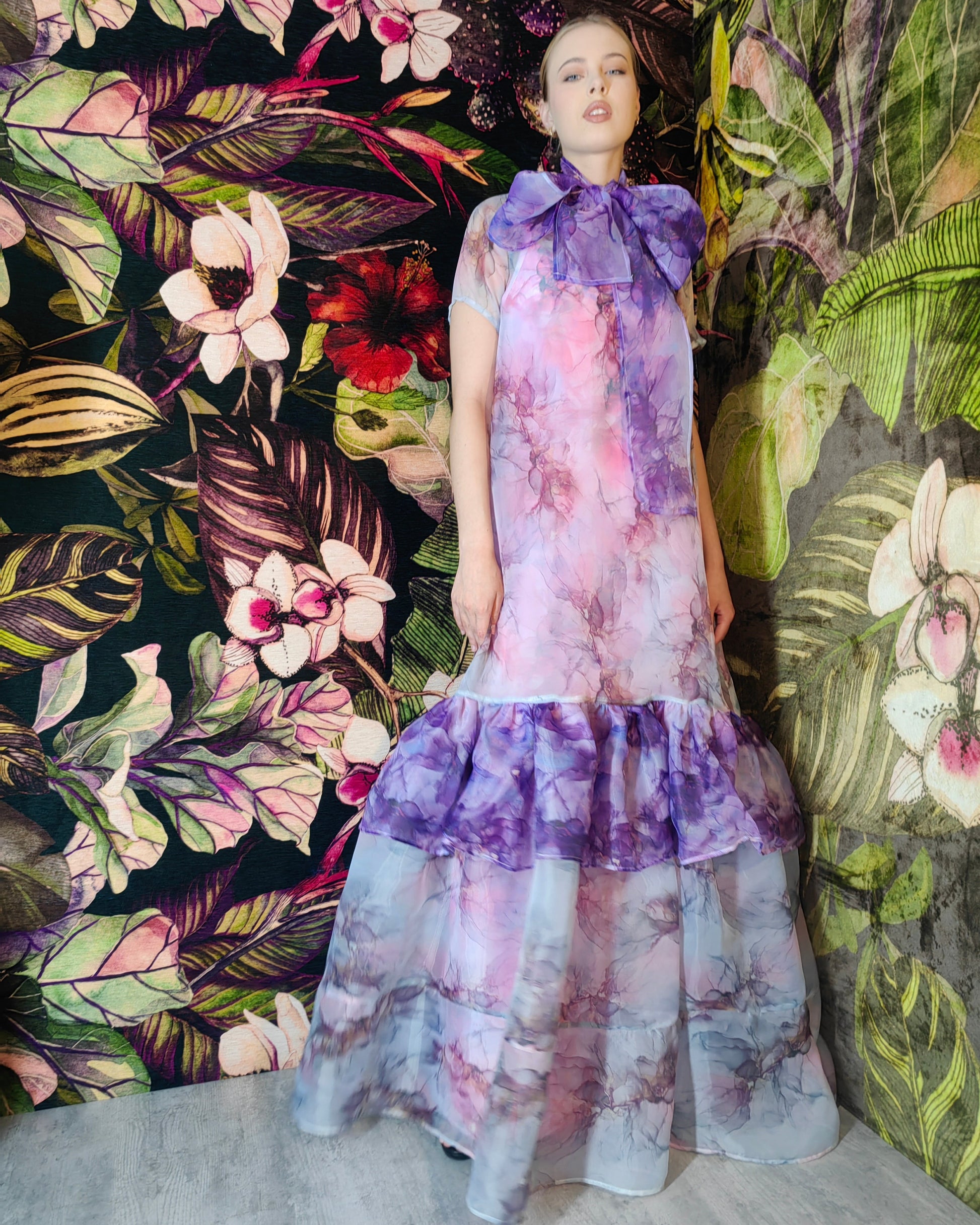 Woman wearing a floral dress standing against a colorful floral and leaf patterned wall.