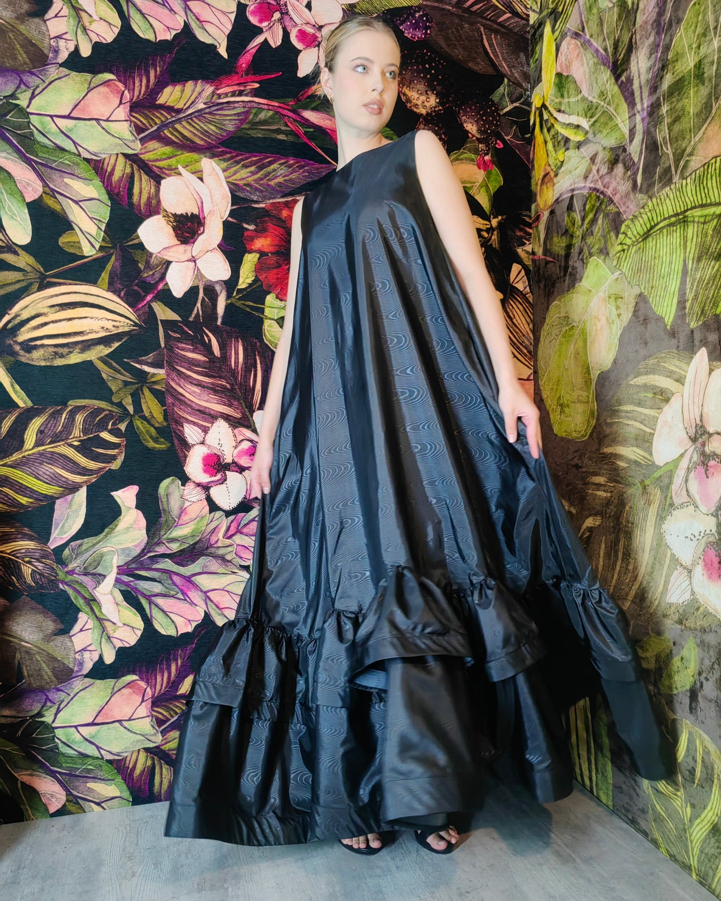 Woman in a black dress standing against a floral and leaf-patterned wall.