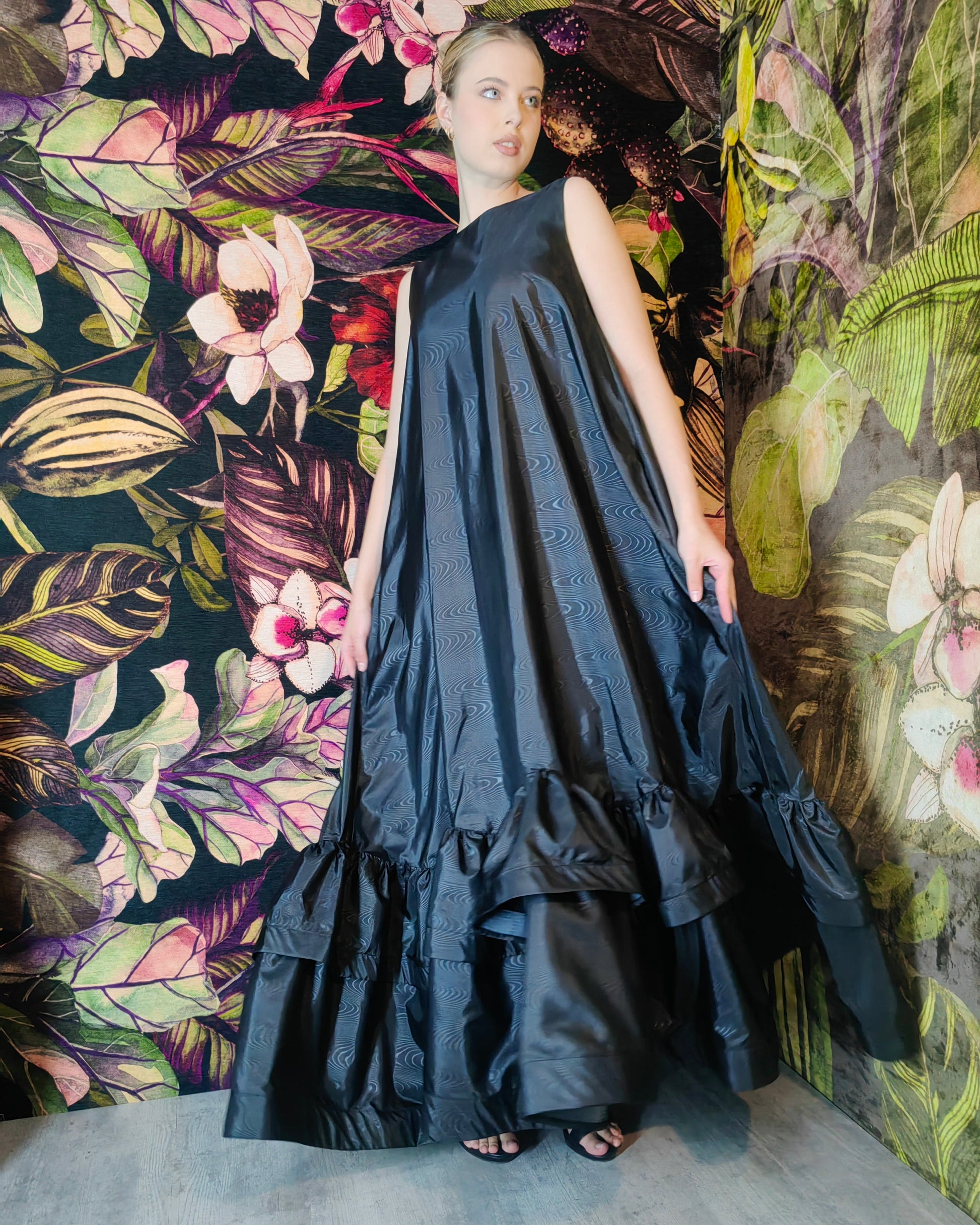 Woman in a black dress standing against a floral and leaf-patterned wall.