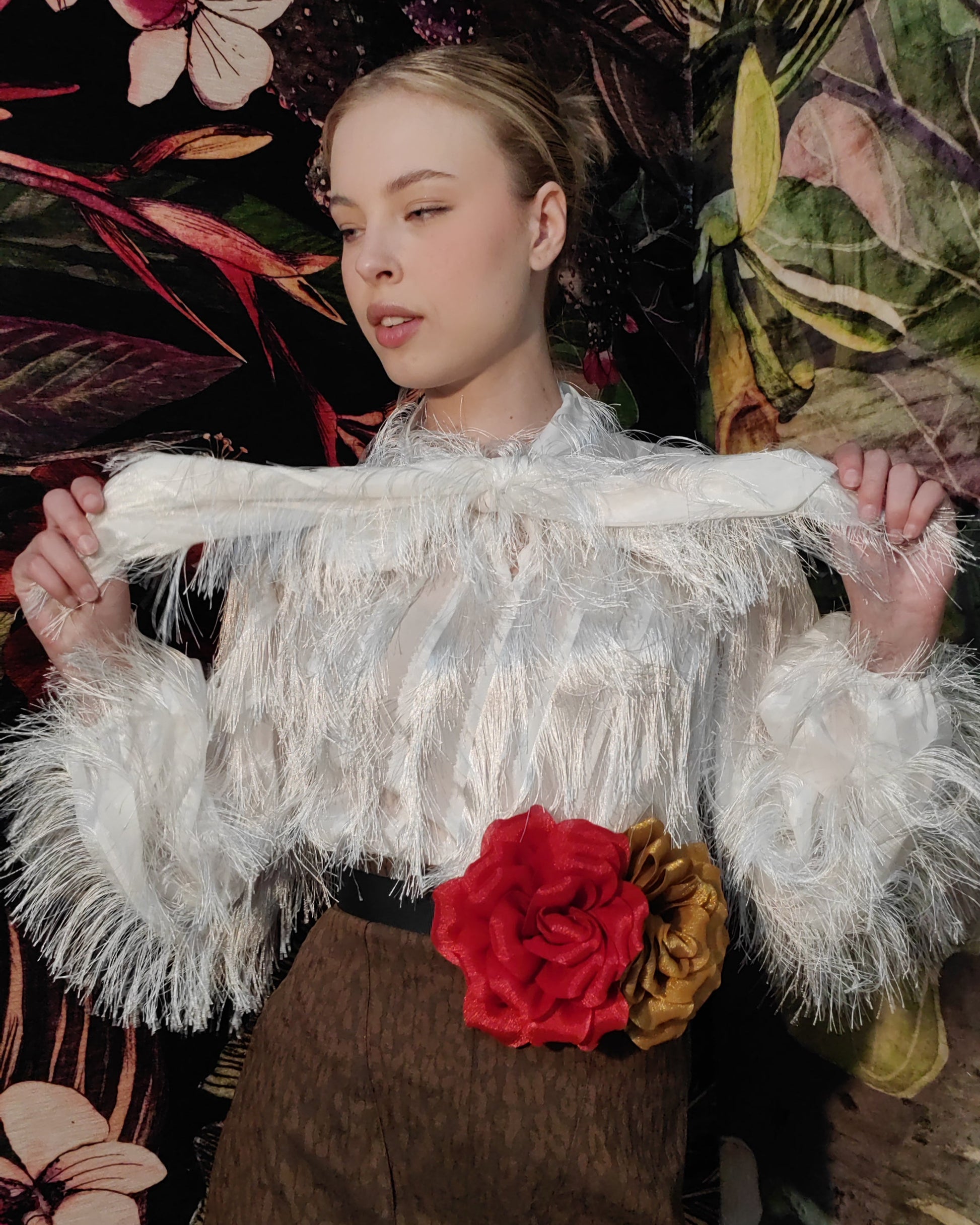 Person wearing a white fringed top with brown overalls against a floral background
